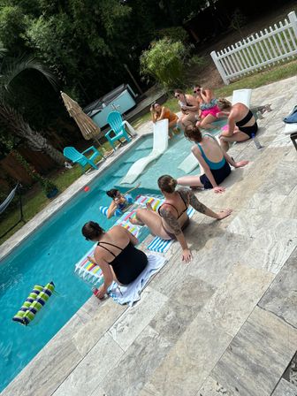 Backyard summer pool party with a group of women lounging on a stone pool deck and on a colorful inflatable, turquoise lounge chairs, hot tub, palm trees and a white picket fence