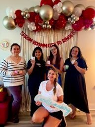 Five friends posing at an indoor bachelorette party in front of a red-and-gold balloon garland and gold fringe backdrop, one kneeling with a bride-to-be sign and others holding party favors.