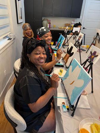 Smiling women at a paint-and-sip night, seated at a long table with easels painting cowboy-hat canvases, holding brushes while enjoying drinks and snacks.