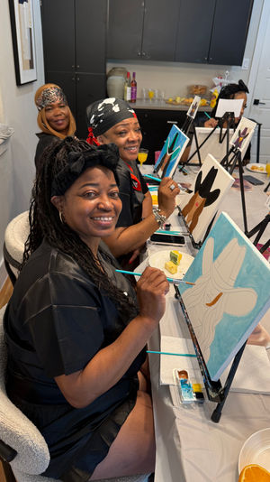 Smiling women at a paint-and-sip night, seated at a long table with easels painting cowboy-hat canvases, holding brushes while enjoying drinks and snacks.