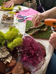 DIY craft scene: hands using a hot glue gun to glue river rocks onto a moss-covered board, surrounded by bright green preserved moss, pink decorative moss, bark pieces, scissors and a tray of pebbles on a tabletop.