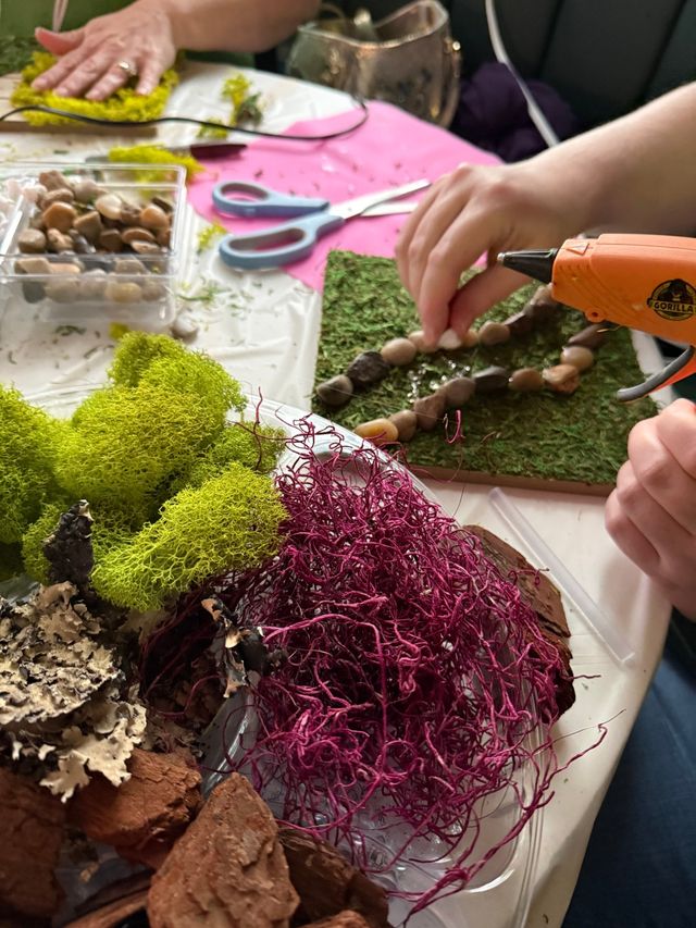 DIY craft scene: hands using a hot glue gun to glue river rocks onto a moss-covered board, surrounded by bright green preserved moss, pink decorative moss, bark pieces, scissors and a tray of pebbles on a tabletop.