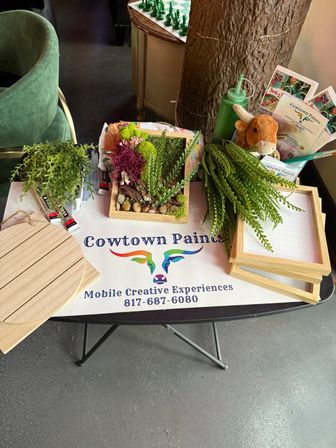Overhead view of a DIY craft workshop table with wooden frames filled with faux moss and tiny succulents, loose fern sprigs, paint tubes, a wooden plaque, brochures and a small stuffed cow toy on a branded tablecloth.