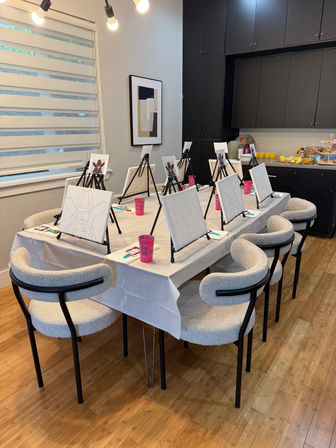 Cozy modern dining room set up for a paint-and-sip party: long white-covered table with tabletop easels holding pre-sketched canvases of a woman in a cowboy hat, pink cups and paint supplies, cushioned chairs, and a buffet of pastries and mimosas against dark cabinets.