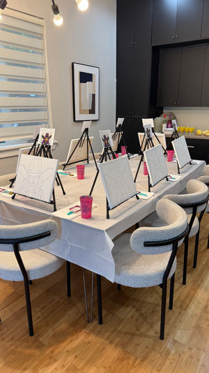 Cozy modern dining room set up for a paint-and-sip party: long white-covered table with tabletop easels holding pre-sketched canvases of a woman in a cowboy hat, pink cups and paint supplies, cushioned chairs, and a buffet of pastries and mimosas against dark cabinets.