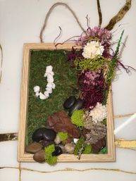 Rustic wooden framed moss wall hanging with preserved green moss, pink and white dried flowers and purple Spanish moss on the right, river rocks, bark and bright green reindeer moss accents, white pebble 'J' detail and twine hanger — indoor wall decor