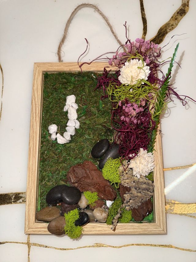 Rustic wooden framed moss wall hanging with preserved green moss, pink and white dried flowers and purple Spanish moss on the right, river rocks, bark and bright green reindeer moss accents, white pebble 'J' detail and twine hanger — indoor wall decor