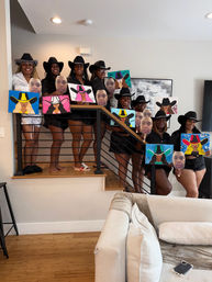 Women wearing cowboy hats pose on a staircase in a modern living room, each holding colorful cowgirl-themed canvas paintings and circular face cutouts from a group painting party.