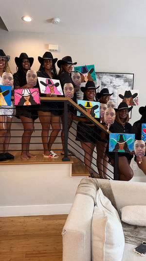 Women wearing cowboy hats pose on a staircase in a modern living room, each holding colorful cowgirl-themed canvas paintings and circular face cutouts from a group painting party.