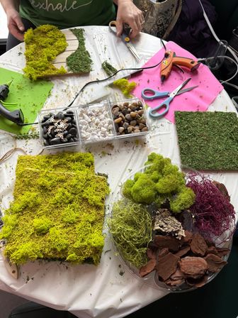 Top-down view of a DIY moss craft workstation with preserved bright green and dark moss panels, loose reindeer moss and bark, a divided tray of black, white and river pebbles, scissors and hot glue guns on a plastic-covered table.