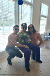 Three people posing in a bright room with large windows overlooking a pool: an older person in a novelty police cap, sunglasses, colorful top and purple pants sits between two shirtless men wearing sunglasses, all smiling and playful.