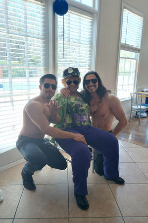 Three people posing in a bright room with large windows overlooking a pool: an older person in a novelty police cap, sunglasses, colorful top and purple pants sits between two shirtless men wearing sunglasses, all smiling and playful.