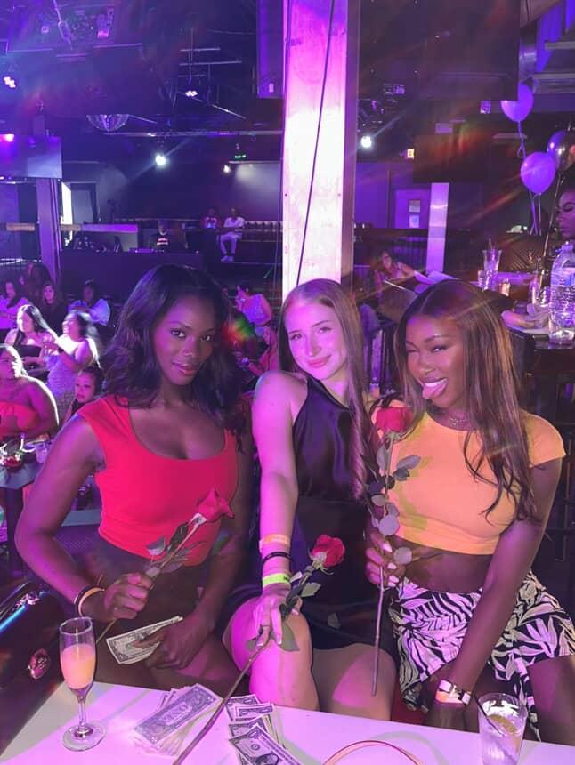 Three women posing at a lively nightclub VIP table under purple lighting, each holding a red rose and cash with cocktails on the table and a crowded dance floor behind them.