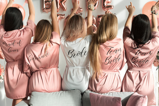 Bachelorette party scene: five women seen from behind in satin robes — bride in white and bridesmaids in pink — arms raised in front of rose-gold balloons and a couch.