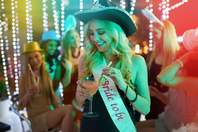 Smiling woman wearing a bride-to-be sash and glittery cowboy hat holding a cocktail at a lively bachelorette party with friends under colorful string lights