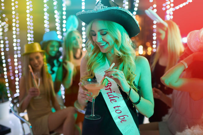 Smiling woman wearing a bride-to-be sash and glittery cowboy hat holding a cocktail at a lively bachelorette party with friends under colorful string lights