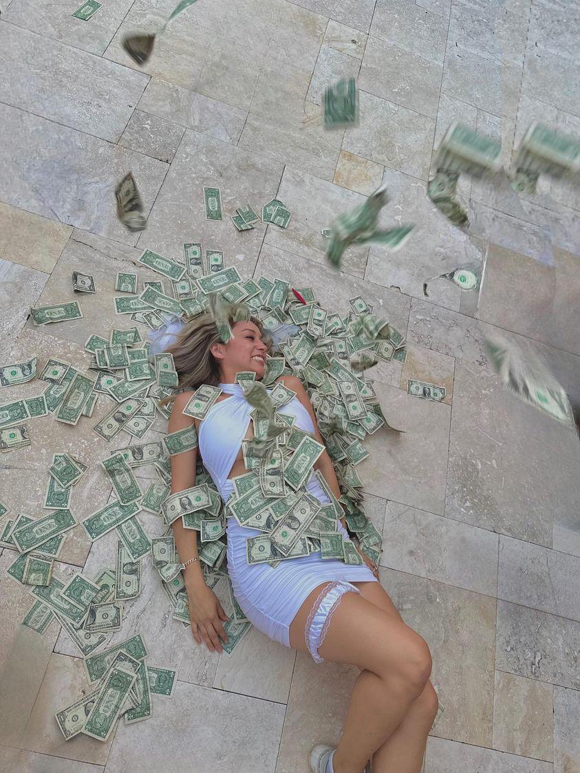 Woman in white dress laughing while lying on light stone tile patio as one-dollar bills rain down and cover her