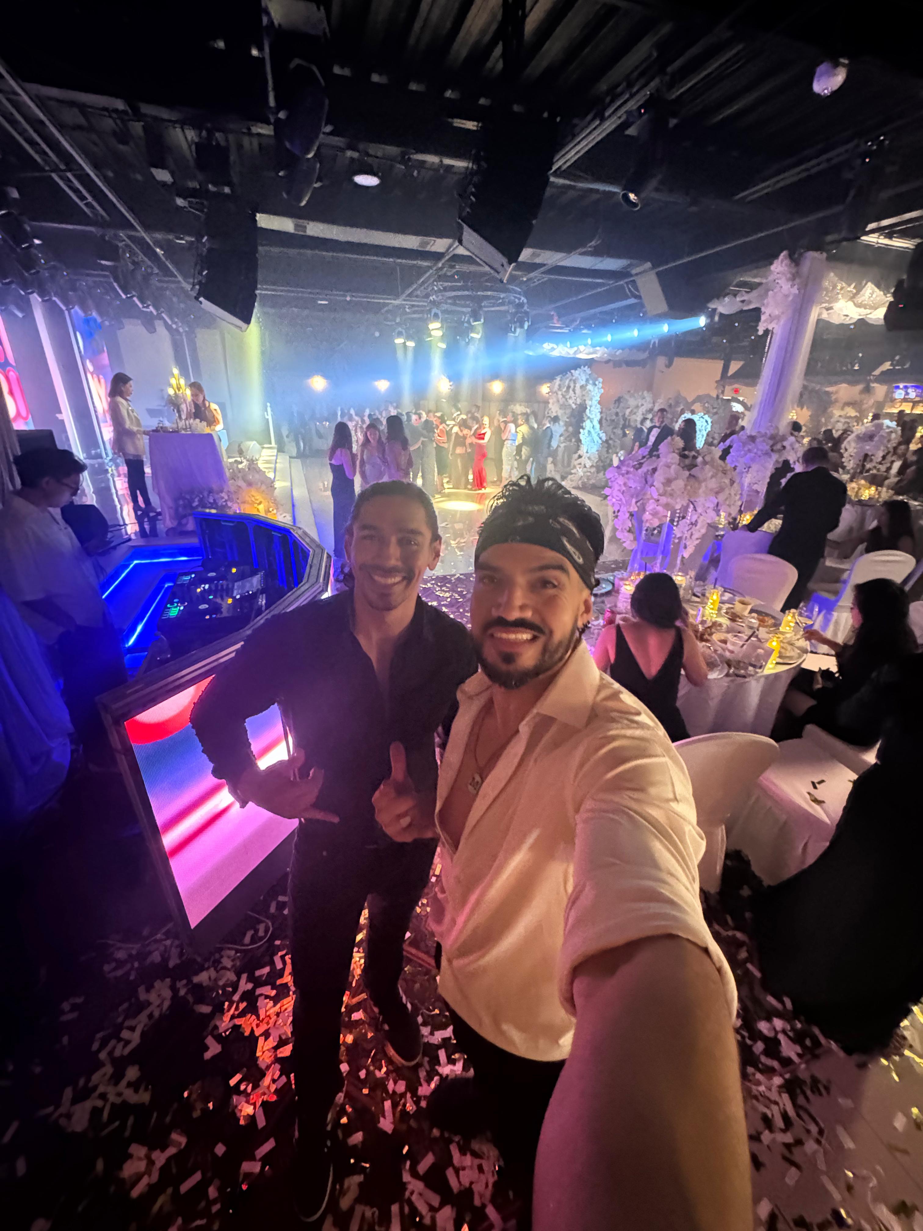 Two men taking a selfie at a lively indoor event venue with a DJ booth and colorful LED lights, confetti-covered floor, crowded dance floor and floral table centerpieces.