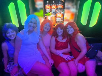 Five friends in colorful wigs and party dresses smiling on a couch in a neon-lit nightclub lounge with green backlighting and a backlit cocktail poster — fun night out group photo