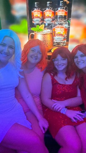 Five friends in colorful wigs and party dresses smiling on a couch in a neon-lit nightclub lounge with green backlighting and a backlit cocktail poster — fun night out group photo