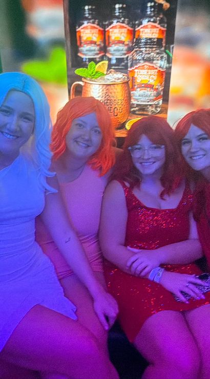 Five friends in colorful wigs and party dresses smiling on a couch in a neon-lit nightclub lounge with green backlighting and a backlit cocktail poster — fun night out group photo