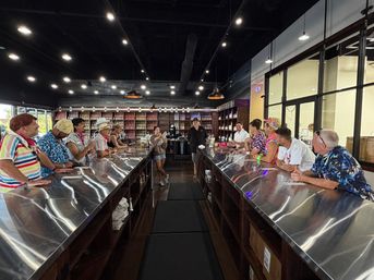 Group of people lined along a long stainless-steel bar in a craft distillery tasting room with shelves of bottles and hanging pendant lights overhead.