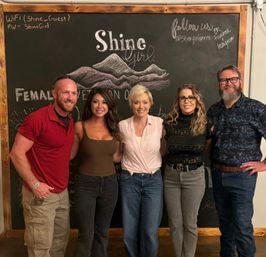 Five smiling adults in casual clothes pose shoulder-to-shoulder for a group photo in front of a large chalkboard wall with a hand-drawn mountain illustration and cursive lettering at an indoor social gathering.