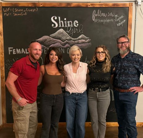 Five smiling adults in casual clothes pose shoulder-to-shoulder for a group photo in front of a large chalkboard wall with a hand-drawn mountain illustration and cursive lettering at an indoor social gathering.