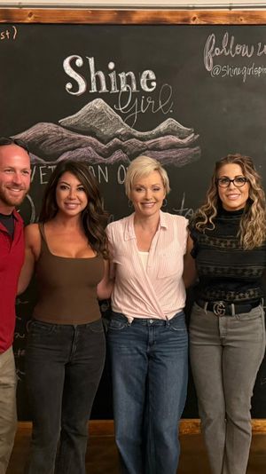Five smiling adults in casual clothes pose shoulder-to-shoulder for a group photo in front of a large chalkboard wall with a hand-drawn mountain illustration and cursive lettering at an indoor social gathering.