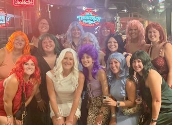 Group of friends in an indoor bar wearing colorful wigs and smiling for a playful group photo beneath neon signs — lively nightlife celebration