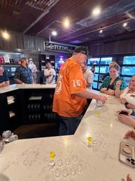 Group at a rustic indoor tasting bar with corrugated metal ceiling; a man in an orange shirt pours sample shots into rows of small glasses on a marble counter while others chat and laugh.