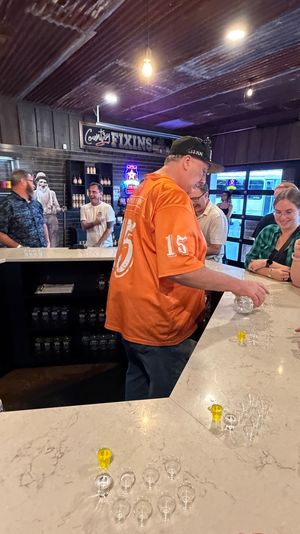 Group at a rustic indoor tasting bar with corrugated metal ceiling; a man in an orange shirt pours sample shots into rows of small glasses on a marble counter while others chat and laugh.