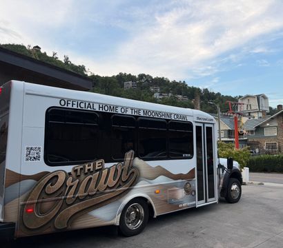White tour shuttle with moonshine-themed graphics parked at a mountain resort base, hillside vacation homes and chairlift towers under a blue sky