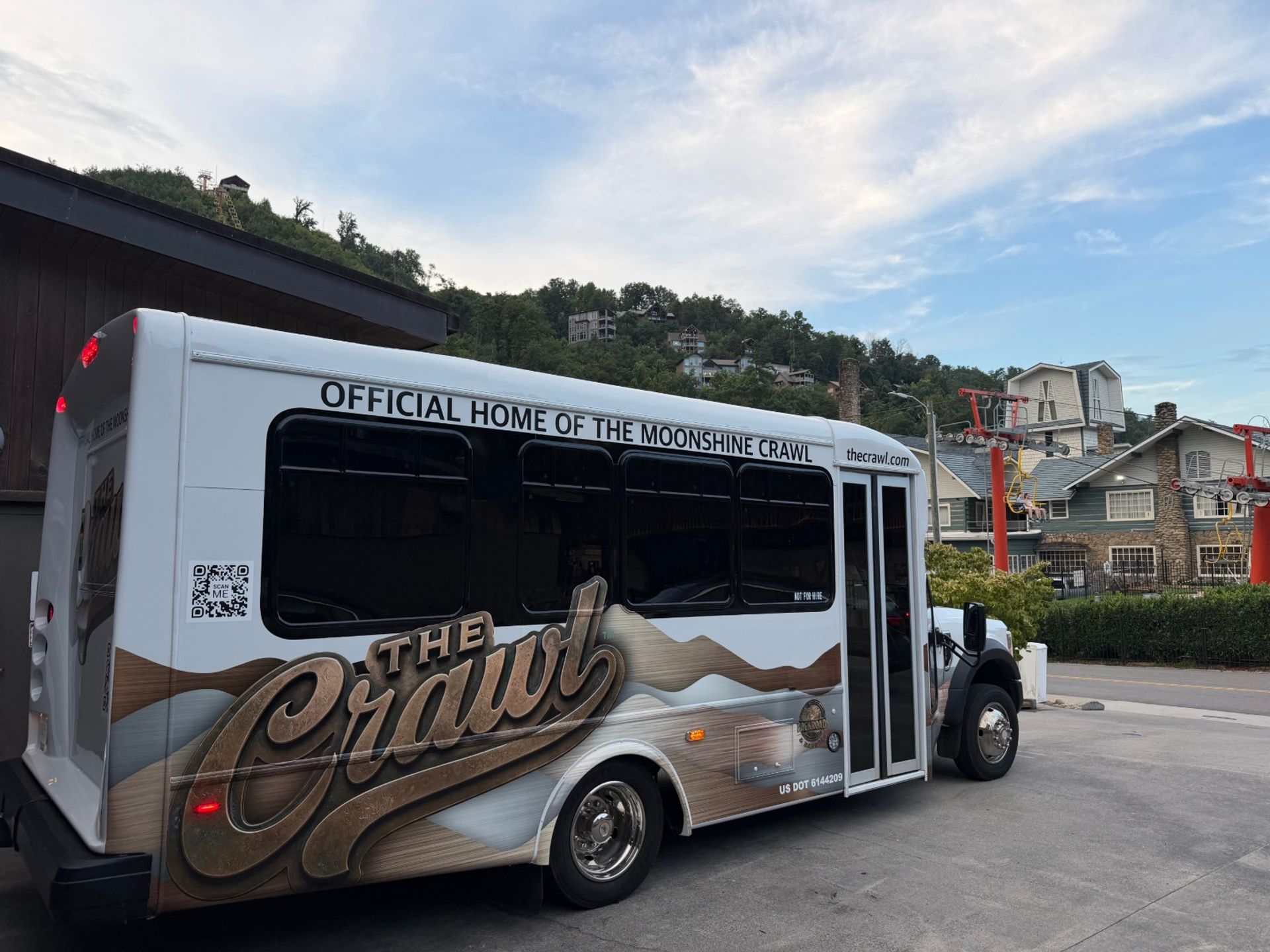 White tour shuttle with moonshine-themed graphics parked at a mountain resort base, hillside vacation homes and chairlift towers under a blue sky