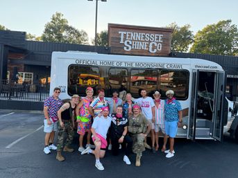 Group of friends in colorful retro outfits and bandanas posing in front of a party shuttle labeled “Official Home of the Moonshine Crawl” parked outside a rustic Tennessee distillery-style storefront