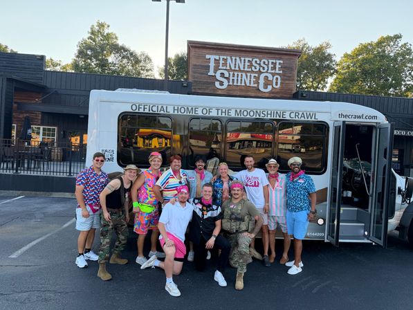 Group of friends in colorful retro outfits and bandanas posing in front of a party shuttle labeled “Official Home of the Moonshine Crawl” parked outside a rustic Tennessee distillery-style storefront