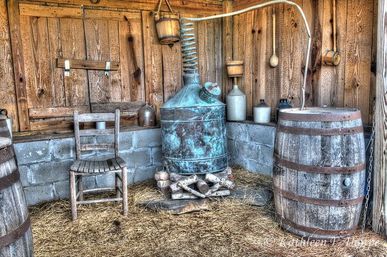 Rustic farm shed interior with a vintage copper distillation still and coil, wooden barrels, clay jugs, a worn chair and straw-covered floor
