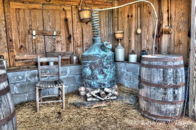 Rustic farm shed interior with a vintage copper distillation still and coil, wooden barrels, clay jugs, a worn chair and straw-covered floor