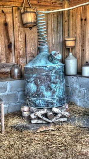 Rustic farm shed interior with a vintage copper distillation still and coil, wooden barrels, clay jugs, a worn chair and straw-covered floor
