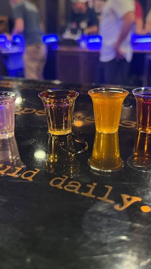 Six vibrant cocktail shot glasses lined up on a glossy bar counter reflecting neon blue lights, with a blurred bar scene and patrons in the background for a lively nightlife vibe