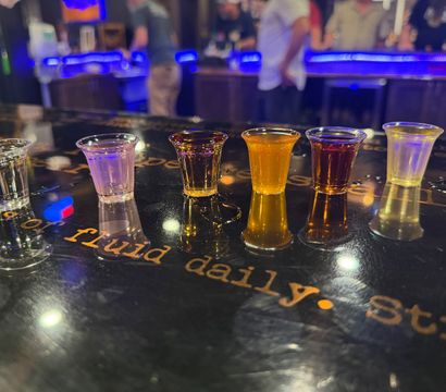 Six vibrant cocktail shot glasses lined up on a glossy bar counter reflecting neon blue lights, with a blurred bar scene and patrons in the background for a lively nightlife vibe
