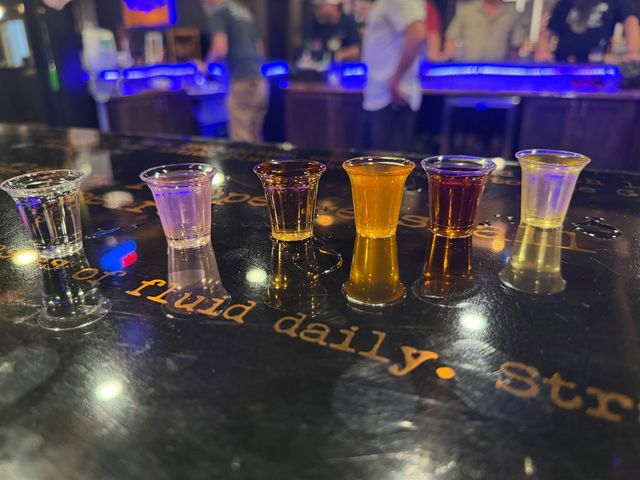 Six vibrant cocktail shot glasses lined up on a glossy bar counter reflecting neon blue lights, with a blurred bar scene and patrons in the background for a lively nightlife vibe