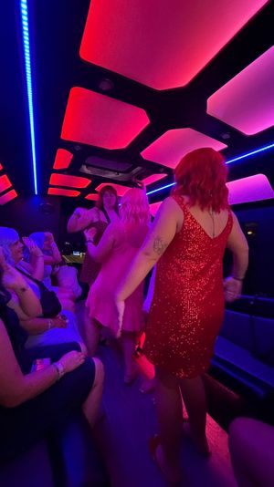 Group of women dancing inside a neon-lit party bus at night, wearing colorful sequined dresses under pink and blue LED ceiling panels