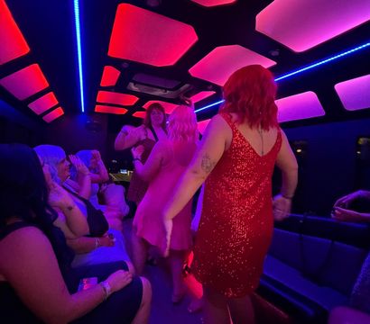 Group of women dancing inside a neon-lit party bus at night, wearing colorful sequined dresses under pink and blue LED ceiling panels