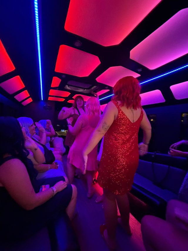 Group of women dancing inside a neon-lit party bus at night, wearing colorful sequined dresses under pink and blue LED ceiling panels