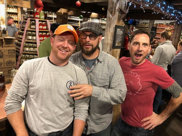 Three men posing and laughing inside a rustic country store with shelves of stacked jars, wooden beams, holiday ornaments and string lights.