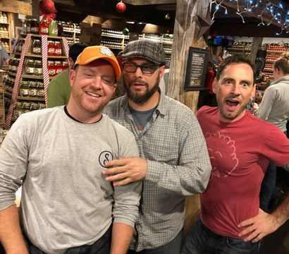 Three men posing and laughing inside a rustic country store with shelves of stacked jars, wooden beams, holiday ornaments and string lights.