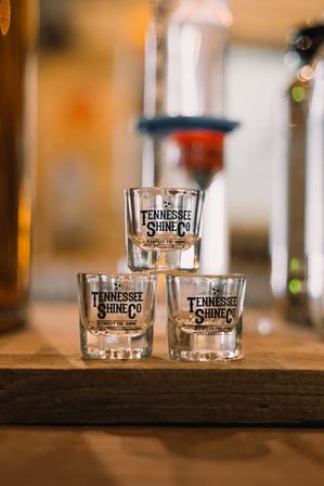 Stacked shot glasses with black logo on a wooden bar in a warm, softly lit Tennessee distillery tasting room.