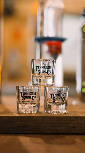 Stacked shot glasses with black logo on a wooden bar in a warm, softly lit Tennessee distillery tasting room.