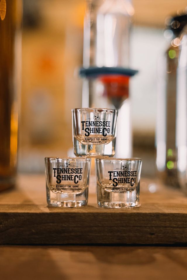 Stacked shot glasses with black logo on a wooden bar in a warm, softly lit Tennessee distillery tasting room.